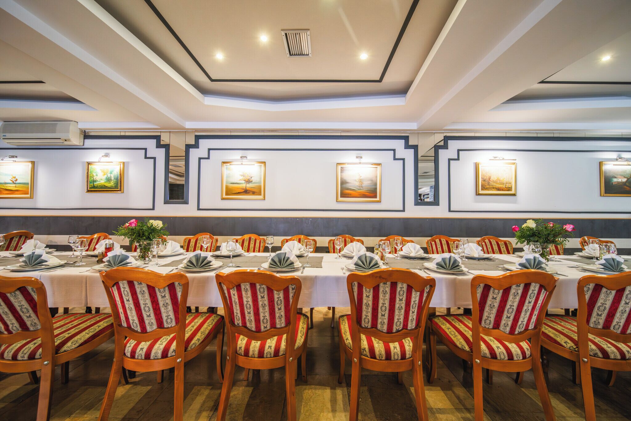 Uma sala de jantar com mesas longas preparadas para o serviço, cadeiras de madeira com estofamento listrado e quadros nas paredes.