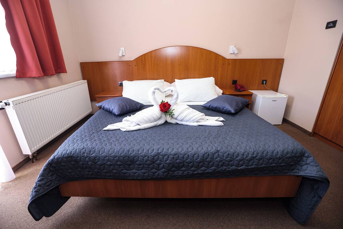 A neatly made bed with a blue quilt, a wooden headboard, and a swan-shaped towel arrangement with a red rose.