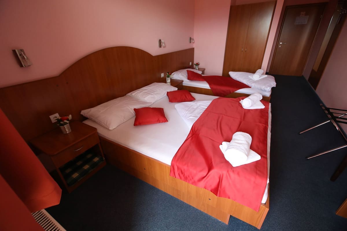 A hotel room with a wooden headboard, a bed with red and white bedding, a small bedside table with a vase, and a wooden wardrobe.