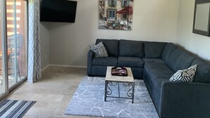 A living room with a dark grey sectional sofa, a small coffee table, a ceiling fan, a flat-screen TV mounted on the wall, and a painting depicting a street scene. - Charming villa on golf course in Old Town Scottsdale (Scottsdale)