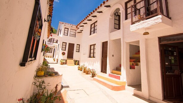 A narrow street with white buildings, balconies, and potted plants. - We are Hostal CasArte Takubamba where you can rest an enjoy! (Sucre, Bolivia)