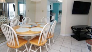 A dining area with a round table and white chairs, a TV on the wall, and a kitchen in the background. - Wall-to-wall ocean views! Dog friendly! Oceanfront balcony with stunning views. (North Myrtle Beach)