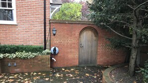 Un mur de briques avec une porte en bois, une borne de recharge pour véhicules électriques fixée au mur, et un espace jardin parsemé de feuilles mortes.