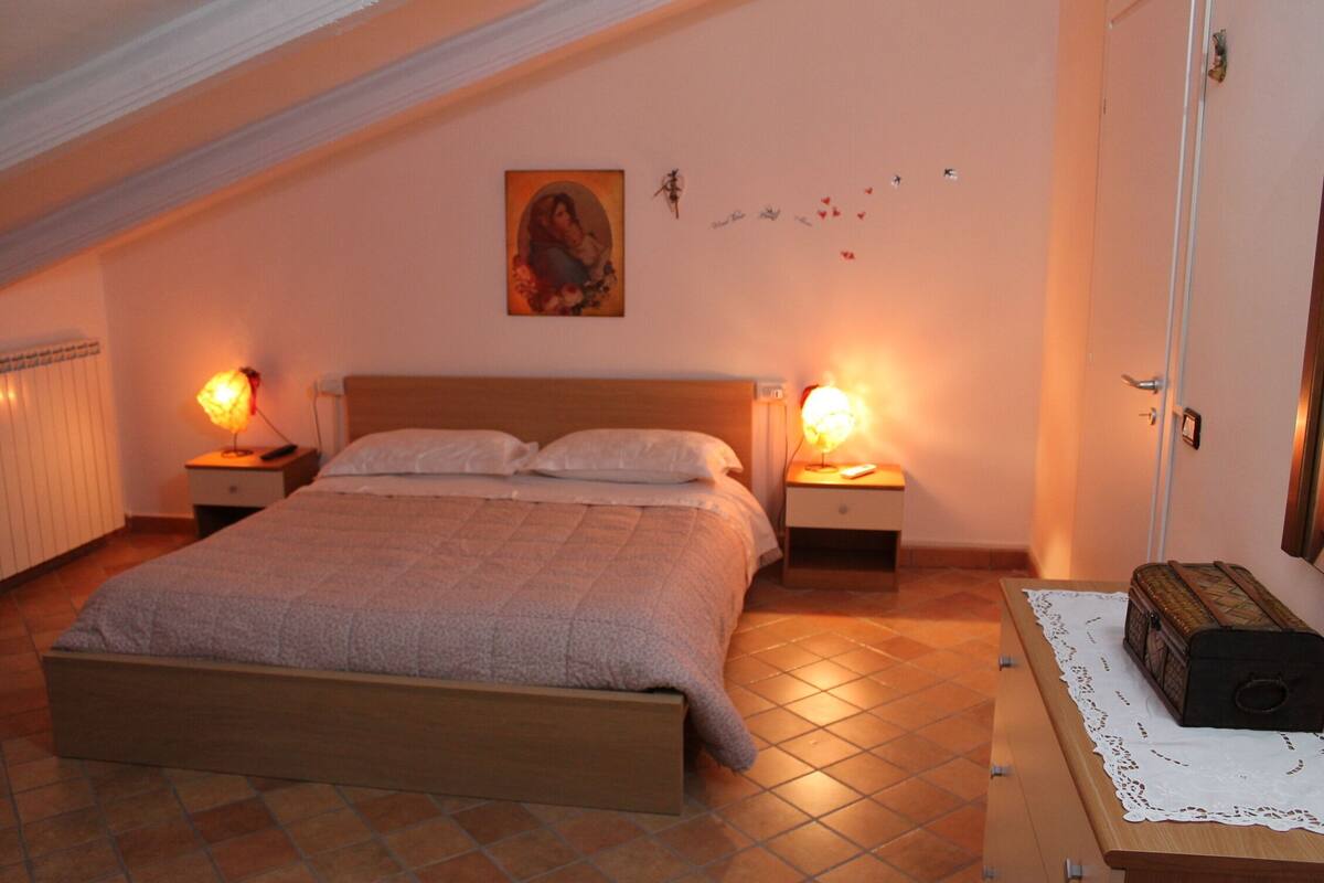 A bedroom with a bed, two bedside tables with lamps, a wooden dresser, and a wall with a picture and small decorations.