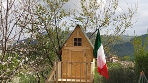 Una casita de juegos de madera con balcón, rodeada de árboles y una bandera.