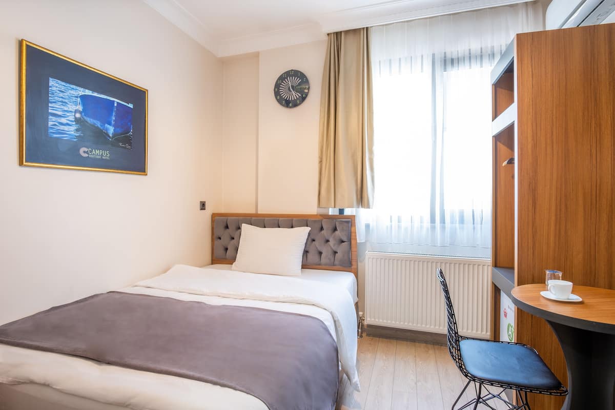 A hotel room with a bed, a chair, a table, a clock, a window with curtains, and a framed picture on the wall.