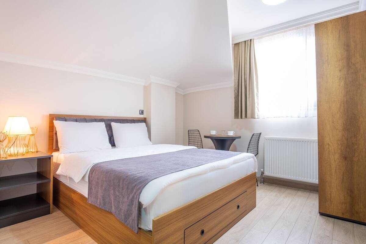 A neatly arranged bedroom with a bed, bedside tables, a small table with cups, a chair, and a window with curtains.