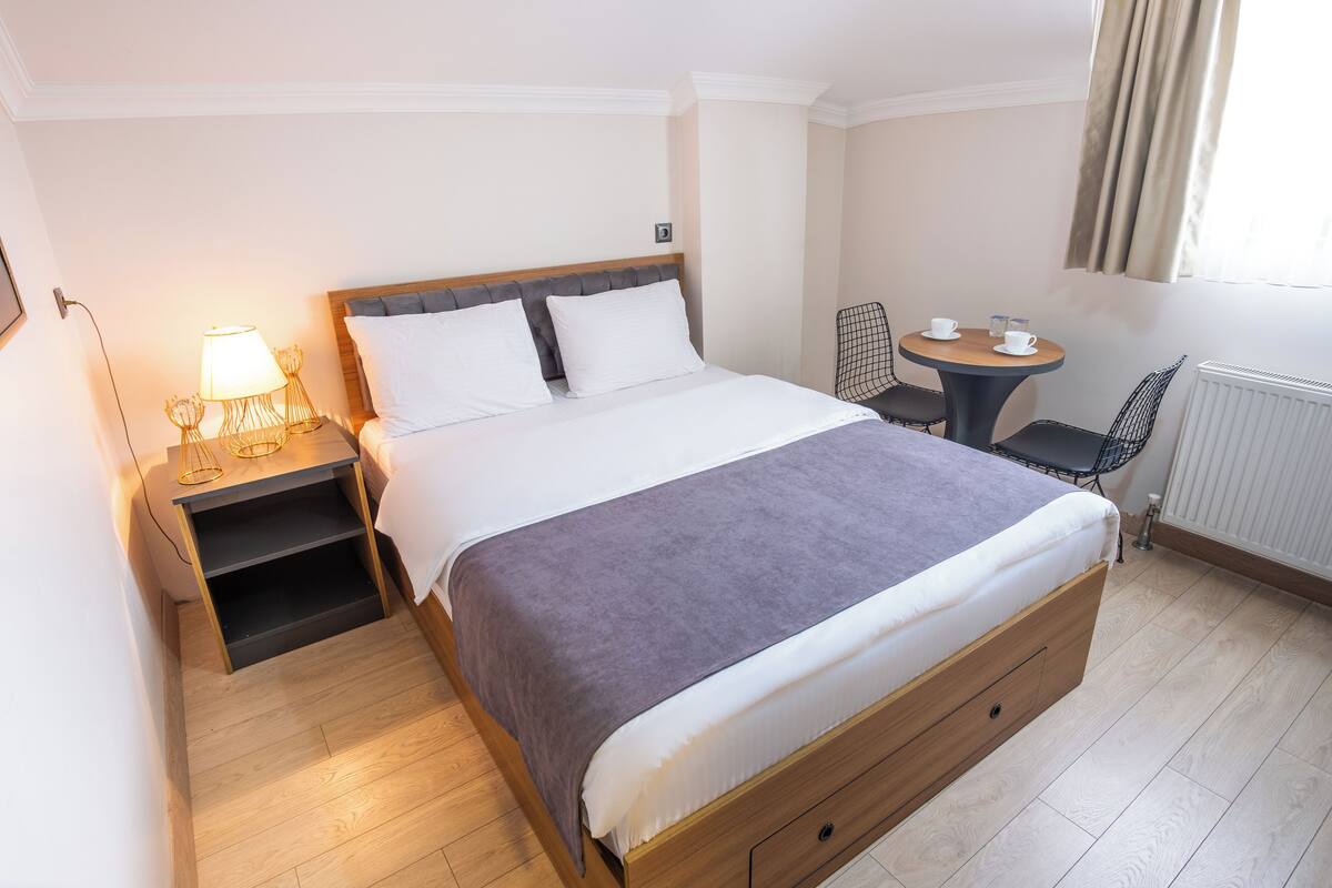 A hotel room with a bed, bedside table, lamp, chair, and a small table with cups.