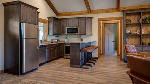 A kitchen with dark wood cabinets, stainless steel appliances, and a wooden ceiling. There are bar stools and a wicker chair in the dining area. - Cozy 2 Bedroom-2 Bath Cottage Near Ski Resorts! Stay at True North Golf Club (Harbor Springs)