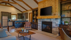 A spacious living area with a fireplace, a television mounted above it, and a kitchen in the background. - Cozy 2 Bedroom-2 Bath Cottage Near Ski Resorts! Stay at True North Golf Club (Harbor Springs)