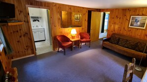 Ein gemütliches Zimmer mit Holzwänden, einer Couch, zwei Sesseln, einem Tisch mit Lampe sowie einer Küchenzeile mit Schränken und Herd.