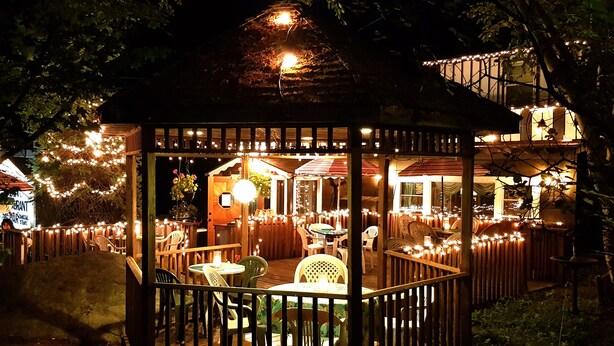 Ein Gazebo mit Holzgeländer, umgeben von Tischen und Stühlen, verziert mit Lichterketten.