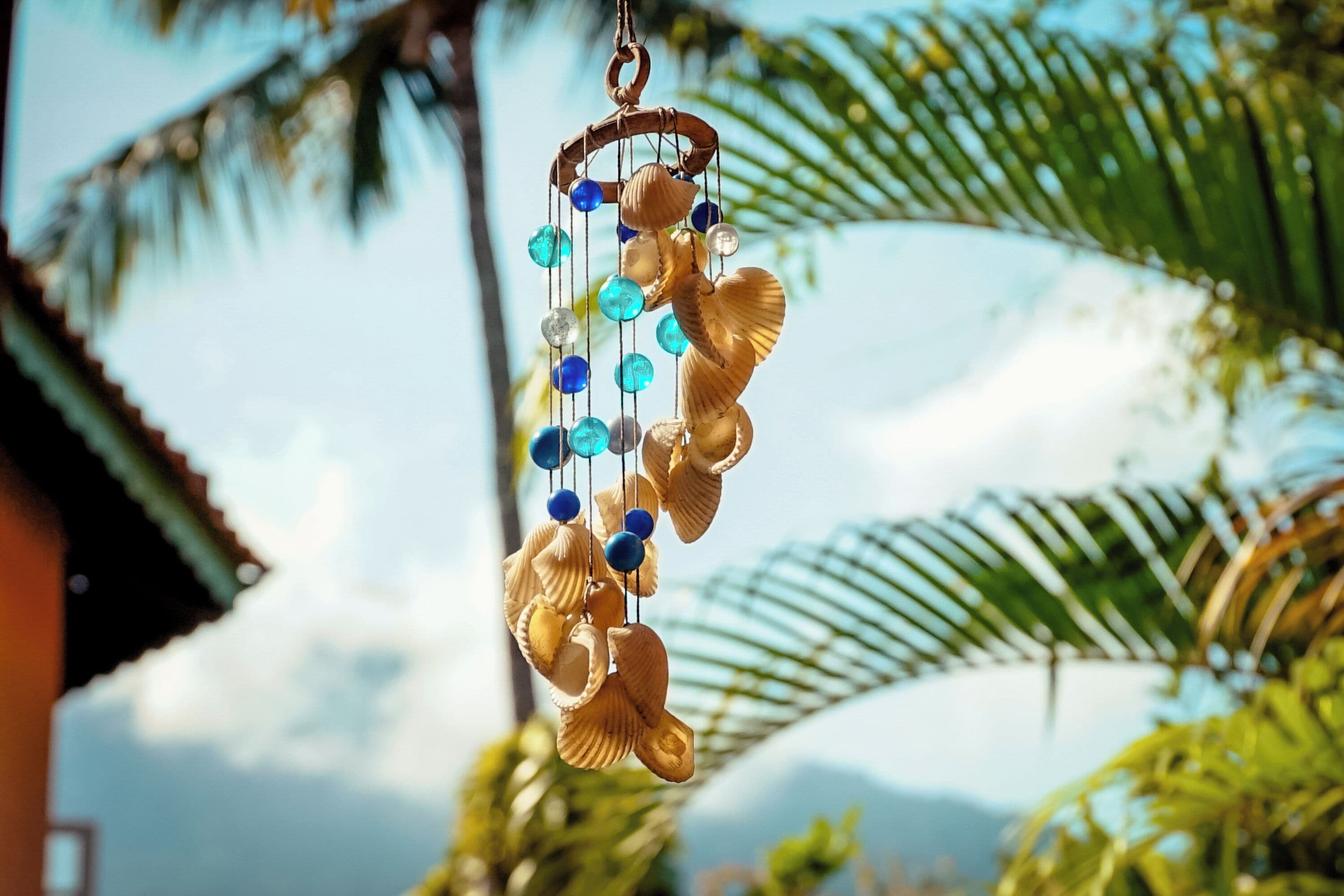 A decorative wind chime with seashells and beads, hanging outdoors with a blurred background of palm trees and a house.