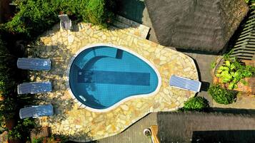 An aerial view of a swimming pool with stone tiles, surrounded by greenery and a stone wall.