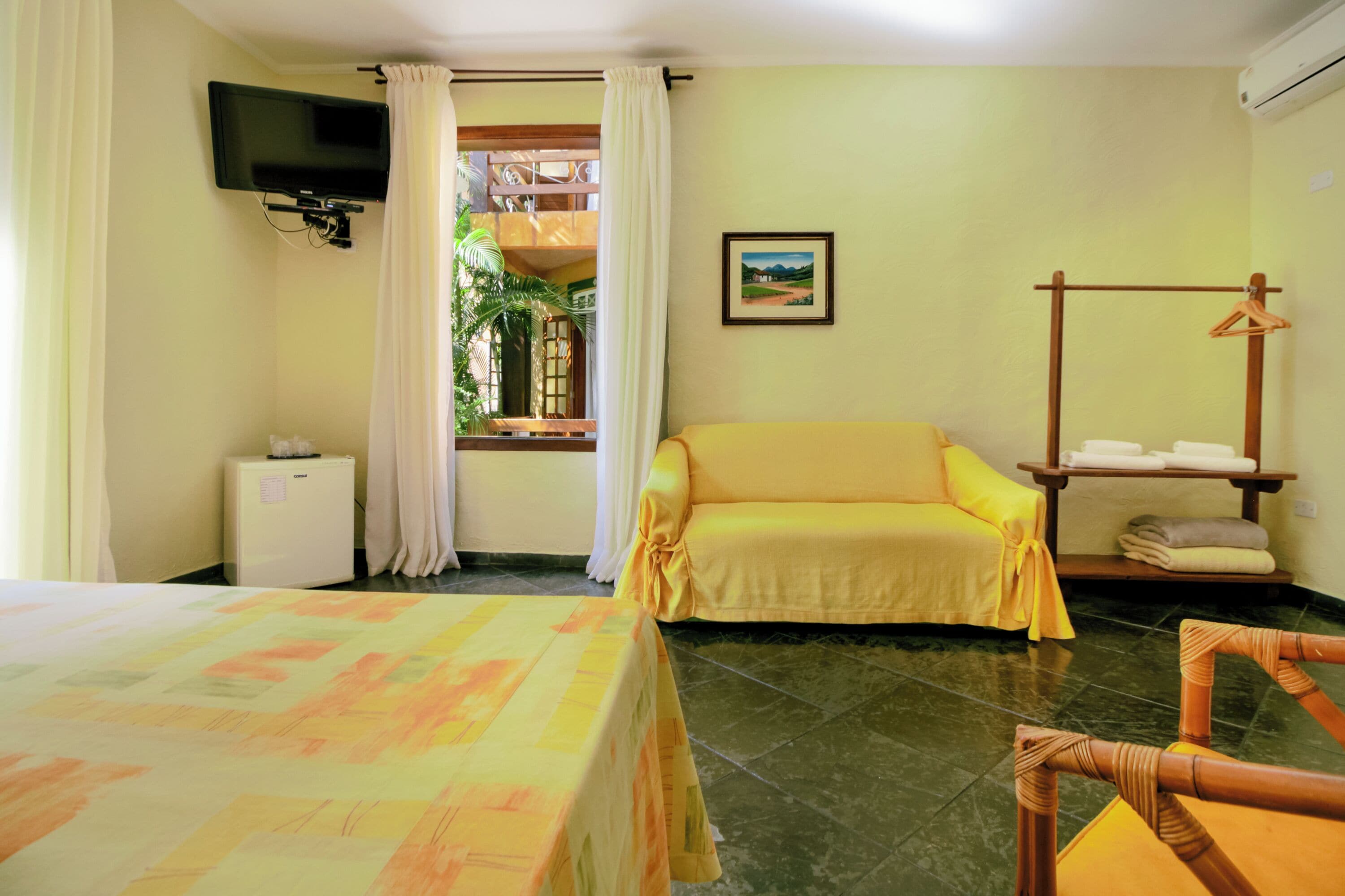 A hotel room with a bed, a sofa, a TV mounted on the wall, a window with curtains, a painting on the wall, and a small table with towels.