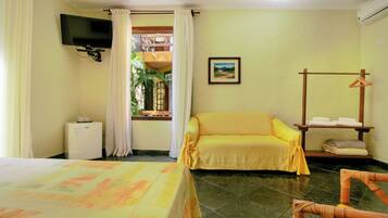 A hotel room with a bed, a sofa, a TV mounted on the wall, a window with curtains, a painting on the wall, and a small table with towels.