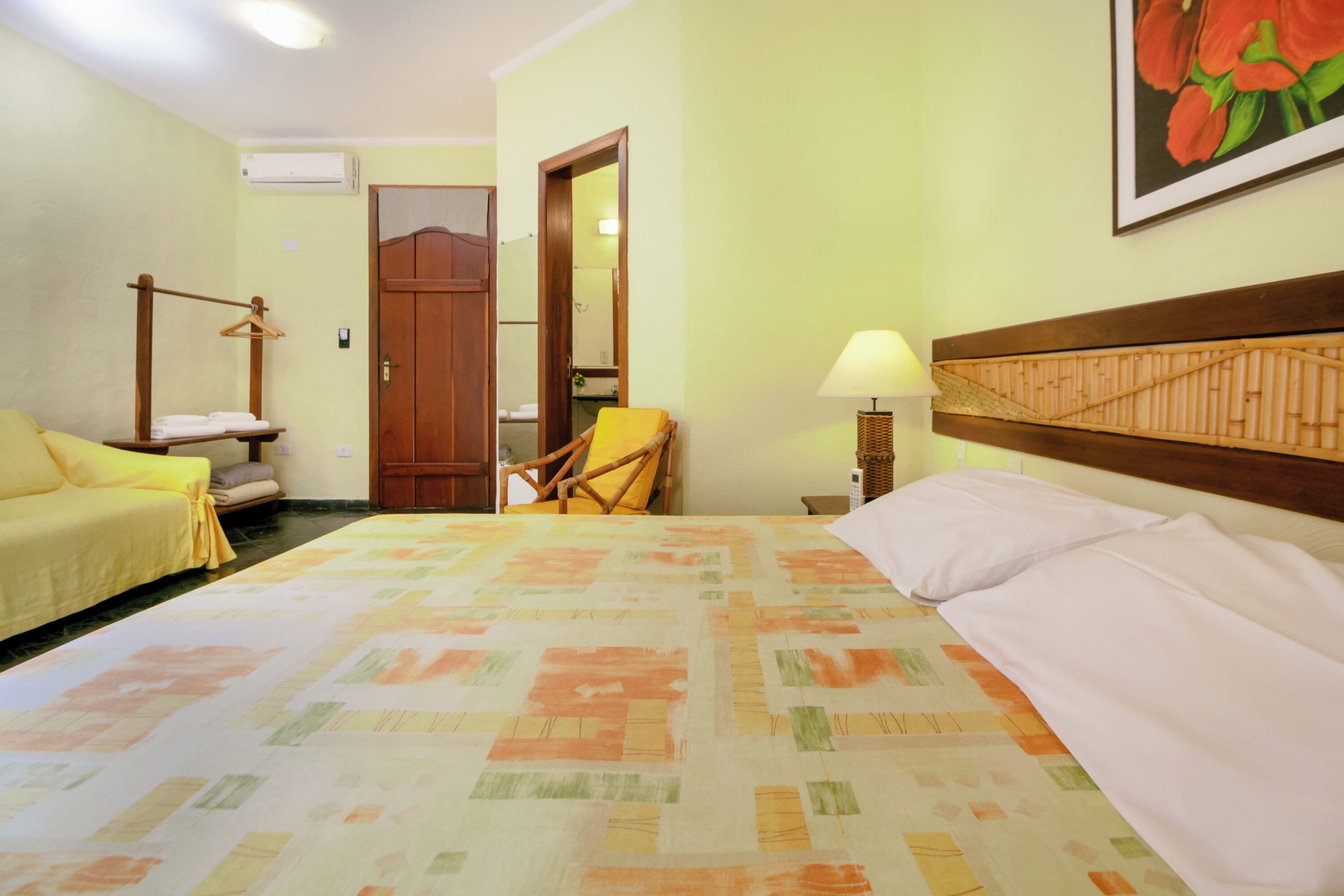 Habitación de hotel con cama, una silla, un sofá, un perchero con toallas y una lámpara, además de un cuadro en la pared.