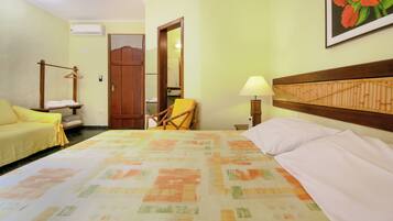 Habitación de hotel con cama, una silla, un sofá, un perchero con toallas y una lámpara, además de un cuadro en la pared.