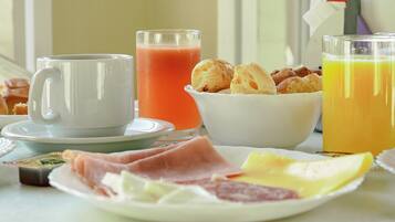 Uma mesa de café da manhã com uma caneca branca, um copo de suco de laranja, uma tigela de croissants e um prato com diversos alimentos.
