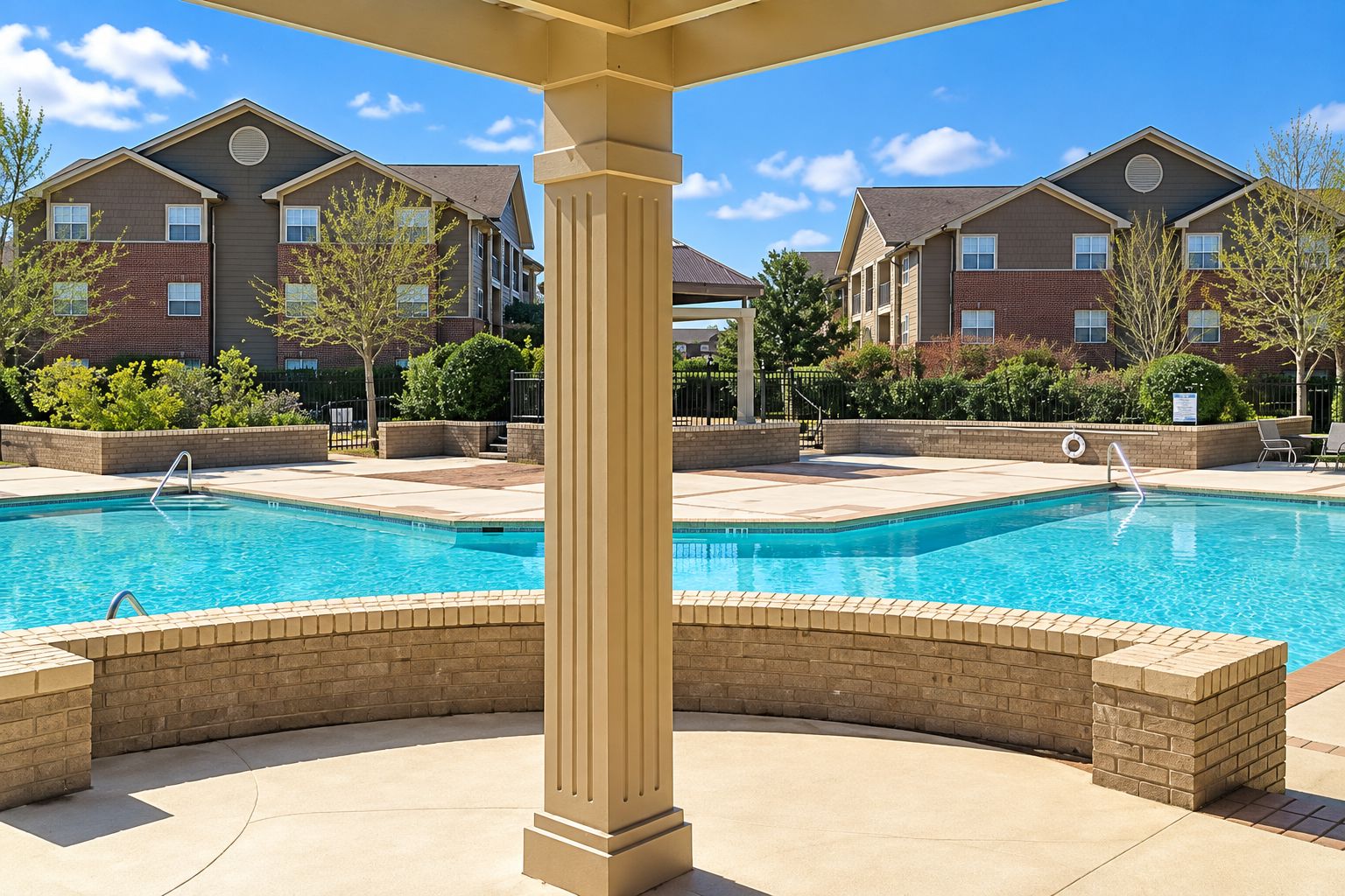 A swimming pool with a beige column in the foreground, surrounded by a brick patio. In the background, there are multi-story buildings with brick facades and windows.