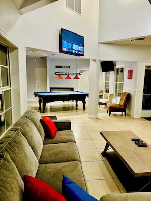 A living room with a pool table, a sofa with red and blue cushions, a wooden coffee table, and a television mounted on the wall.
