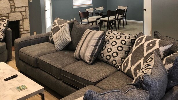 Un salon avec un canapé gris, des coussins à motifs, un mur en pierre et un coin repas comprenant des chaises et une table.