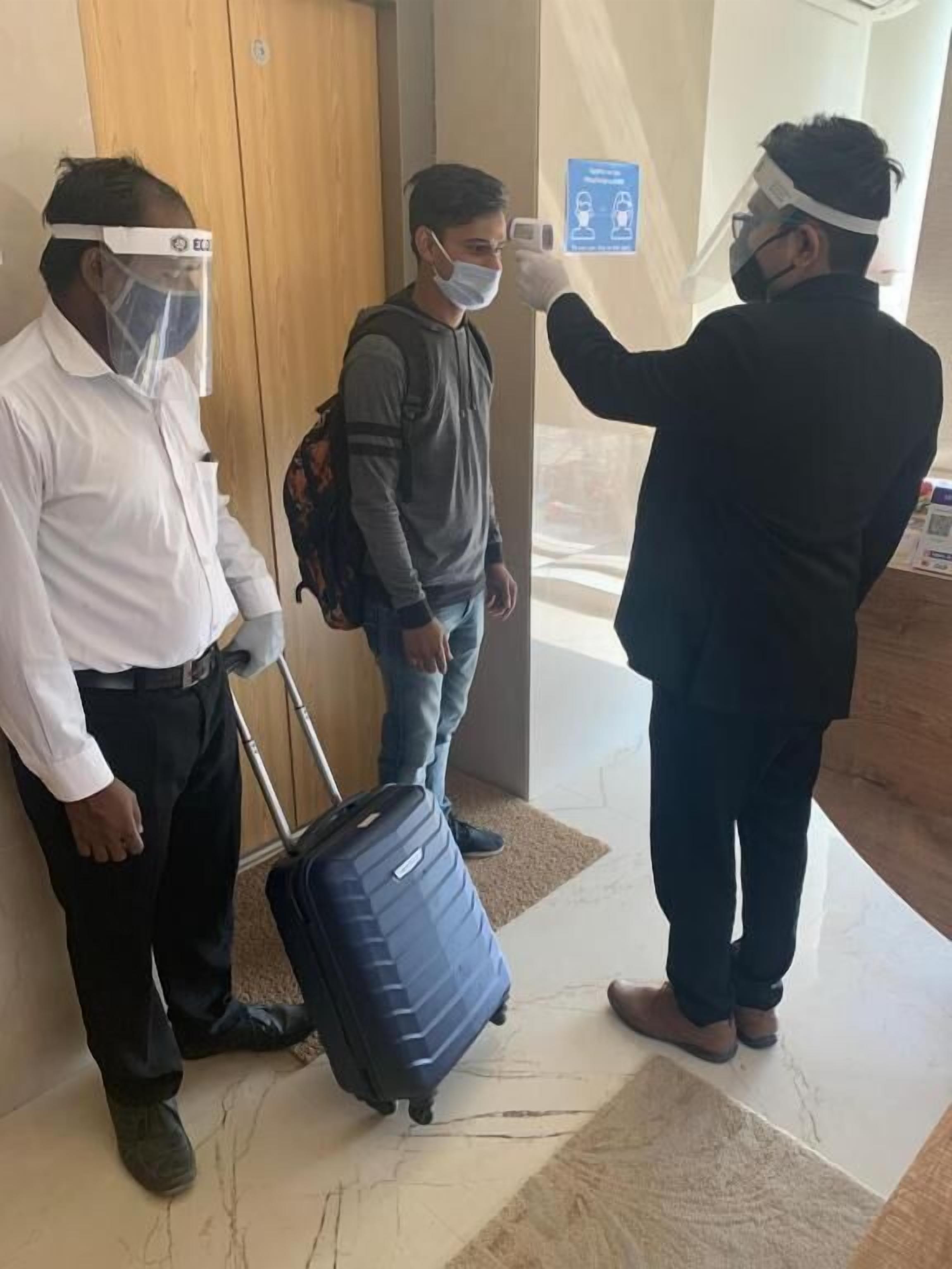 Un'area reception di un albergo con una porta in legno, una valigia blu e una persona che indossa una visiera protettiva.