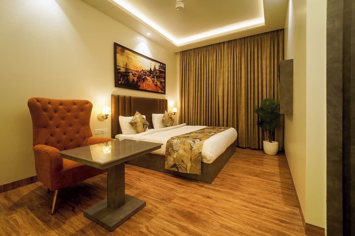 A hotel room with a bed, a tufted armchair, a table, a painting on the wall, and a large window with curtains.