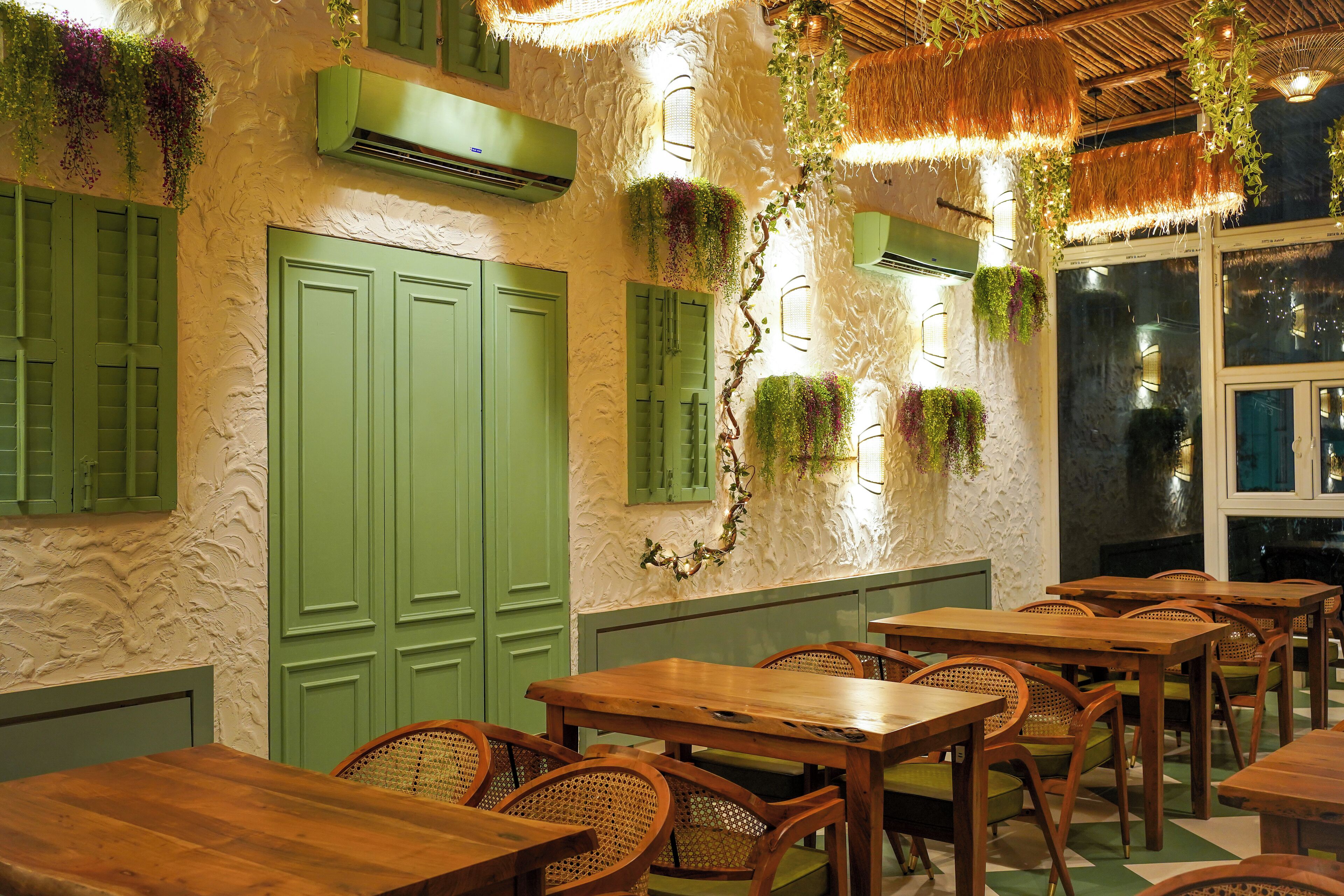 Interior de restaurante con mesas y sillas de madera, contraventanas verdes y plantas colgantes.