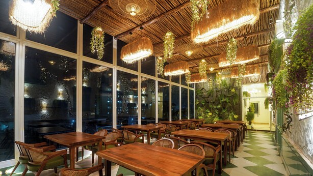 A restaurant with wooden tables and chairs, hanging lights, and large windows. - Sugar Stars Inn (Varanasi)