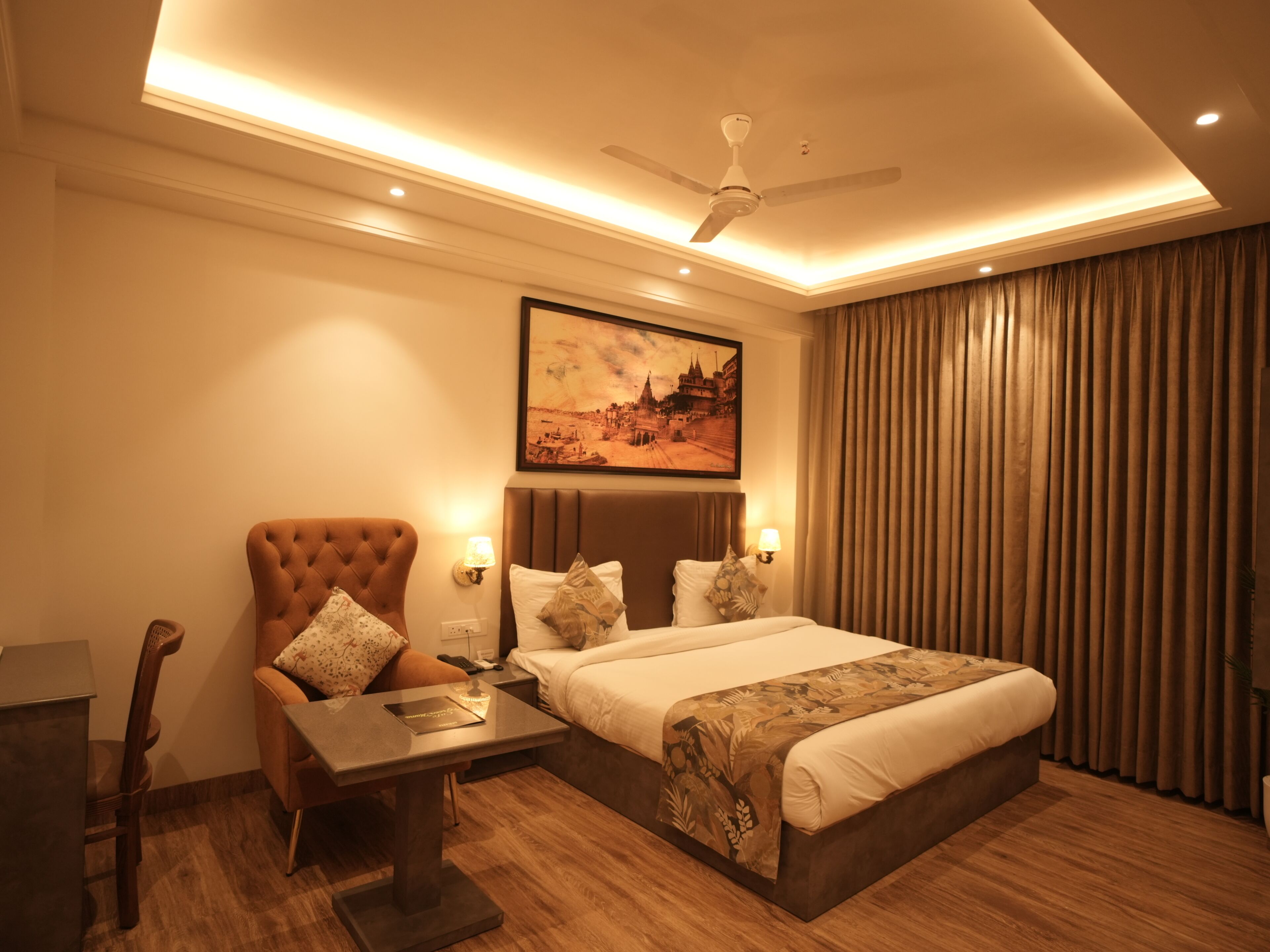 A hotel room with a bed, a desk, a chair, a ceiling fan, and a painting on the wall.
