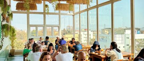 Un restaurant doté de grandes fenêtres, de plantes suspendues et d’un plafond en chaume.