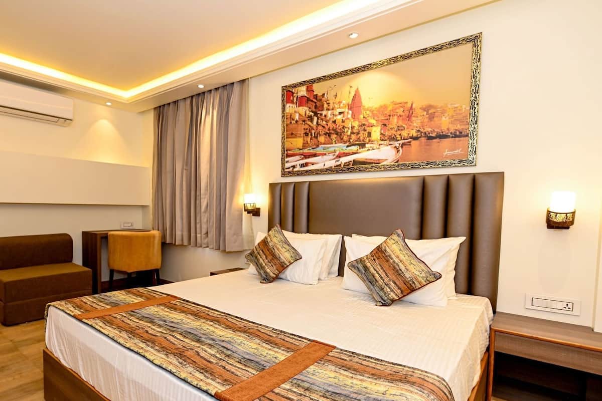 A hotel room with a large bed, a sofa, a chair, a painting, and an air conditioning unit.