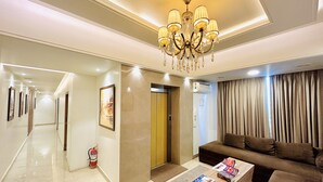 A hotel lobby with a chandelier, a sofa, a coffee table with magazines, a fire extinguisher, and a hallway with framed pictures. - Sugar Stars Inn (Varanasi)
