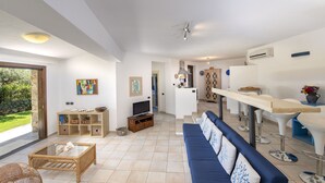 A spacious living area with a blue sofa, a glass coffee table, and a television. There is a wooden cabinet with baskets and a wicker chair. A kitchen area with a bar and stools is visible in the background.