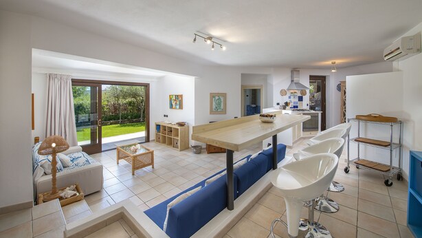 A modern kitchen with a large island, a dining area with a long table, and a seating area with blue cushions.