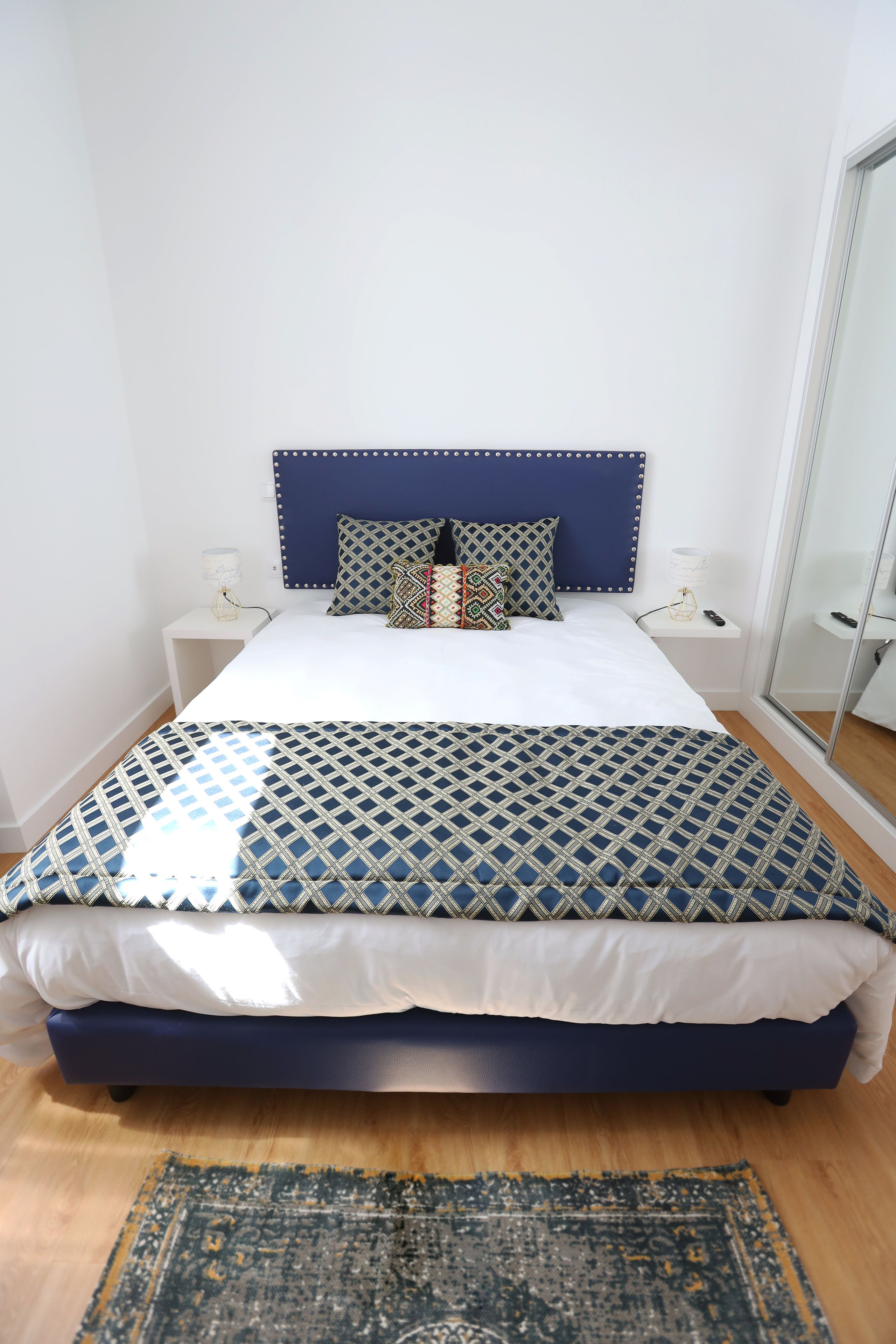 A neatly made bed with a patterned bedspread, a blue headboard, and a bedside table with a lamp.