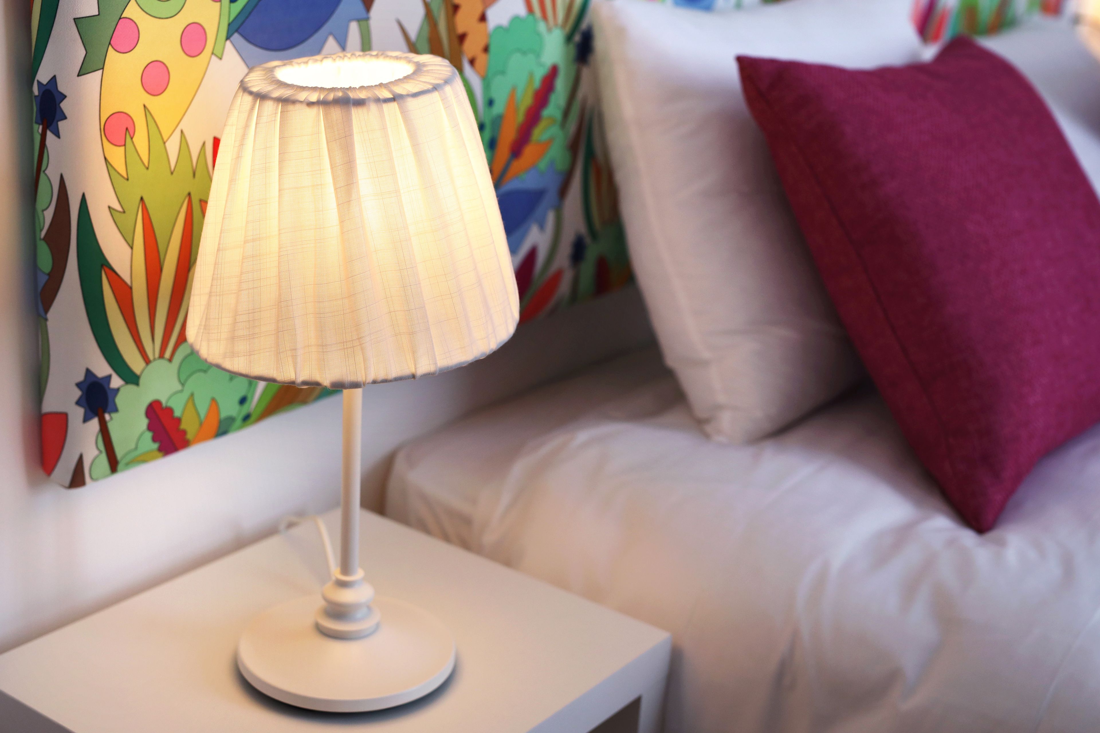 A bedside table with a lamp, a white pillow, and a colorful wall mural.