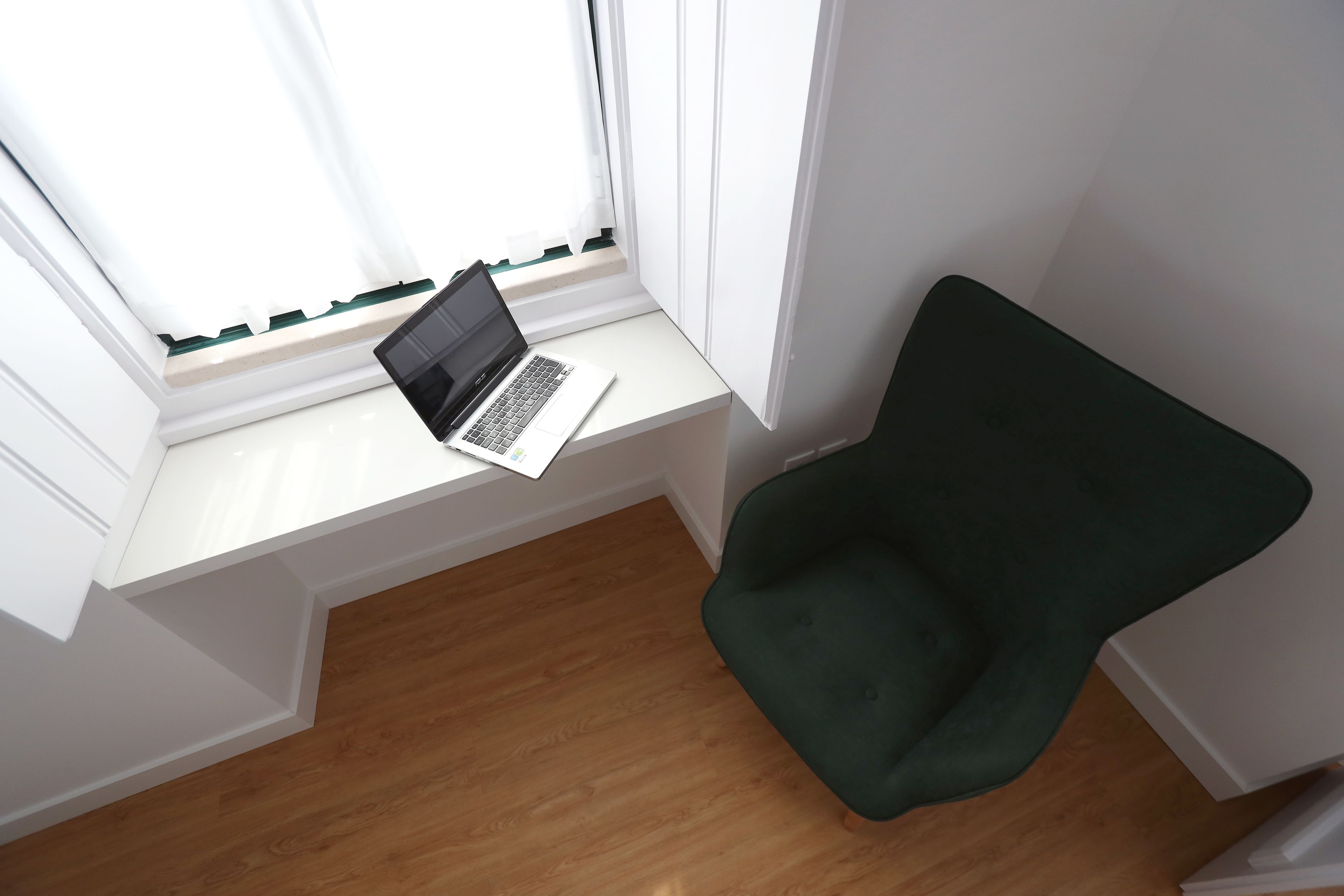 Une pièce avec un ordinateur portable posé sur un bureau blanc près d’une fenêtre, une chaise verte et un plancher en bois.