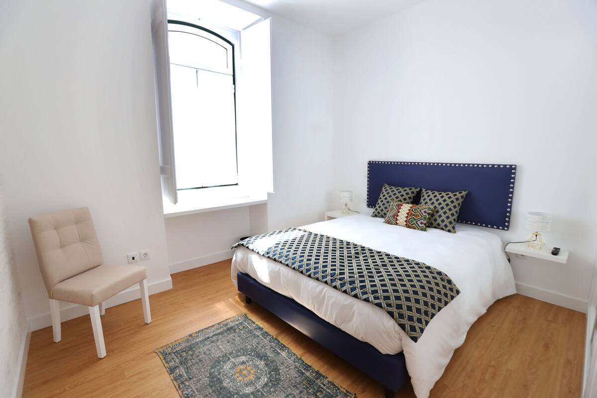Un dormitorio con cama, una silla, una ventana y una mesita de noche.