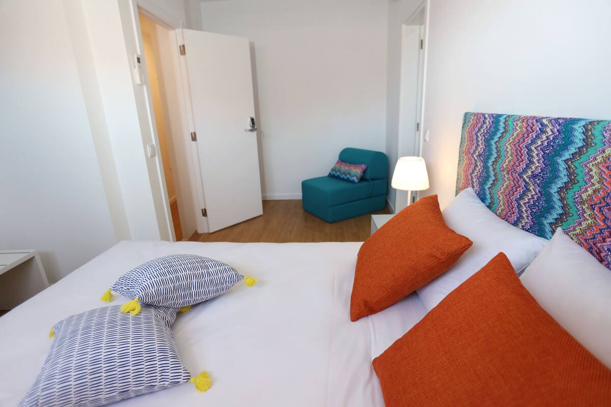 Una habitación de hotel con cama, una silla y un cabecero colorido.