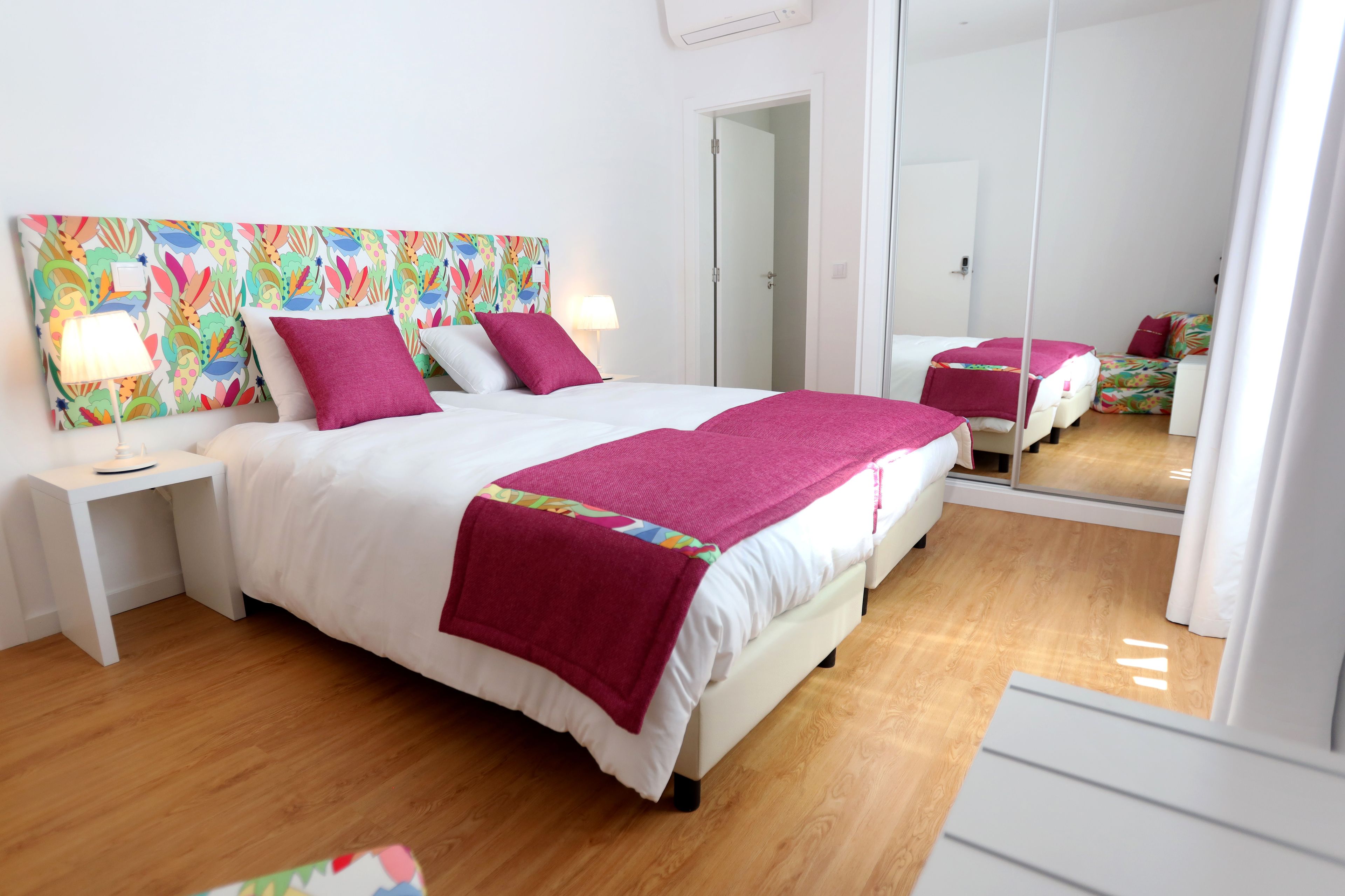 Ein ordentlich eingerichtetes Schlafzimmer mit einem großen Bett, einem Nachttisch, einem Spiegel und einem farbenfrohen Kopfteil.