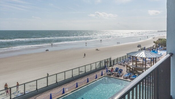 A beach view from a balcony with a pool and lounge chairs.