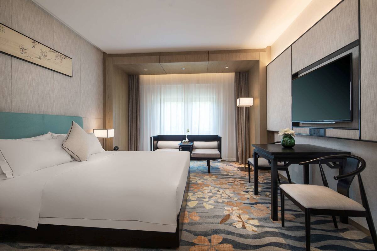 Ein Hotelzimmer mit einem großen Bett, einem Schreibtisch mit Stuhl, einem Fernseher und einem Fenster mit Vorhängen.