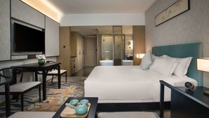 A modern hotel room with a large bed, a desk with a flat-screen TV, a dining table with chairs, and a bathroom visible through a glass door. - Ramada by Wyndham Zhenjiang City Center (Zhenjiang)