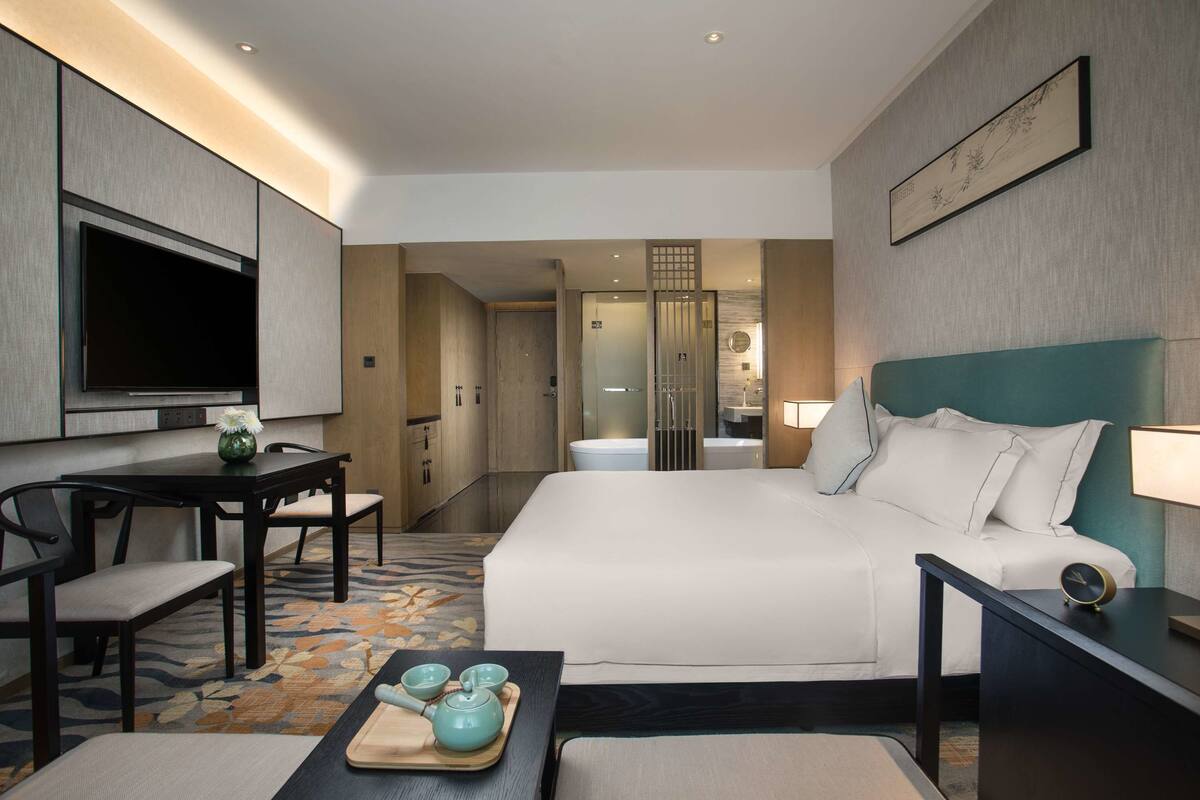 Ein modernes Hotelzimmer mit einem großen Bett, einem Schreibtisch mit Flachbildfernseher, einem Esstisch mit Stühlen und einem durch eine Glastür sichtbaren Badezimmer.