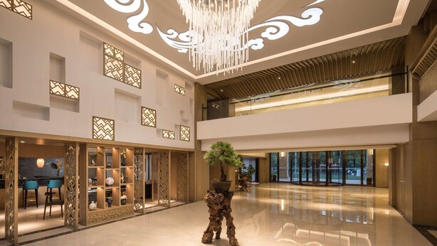 A spacious lobby with a modern chandelier, decorative wall panels, and a central potted tree. - Ramada by Wyndham Zhenjiang City Center (Zhenjiang)