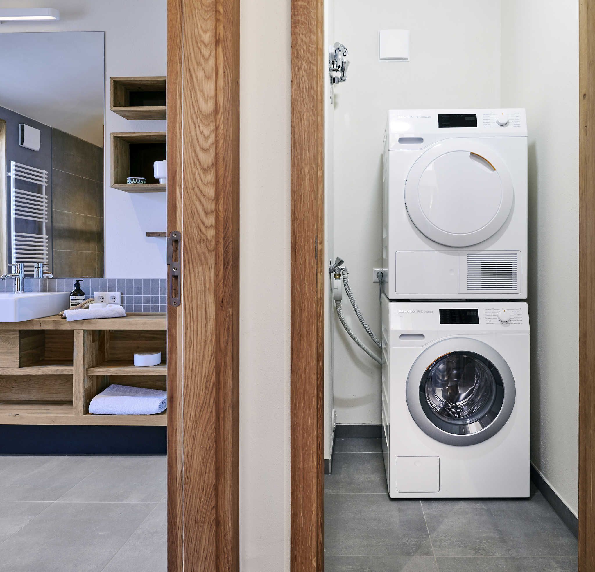 A compact laundry room with a stacked washer and dryer, a wooden shelf with towels, and a modern bathroom with a sink and mirror.