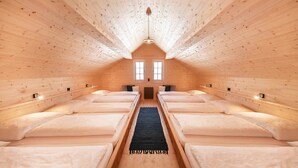 Habitación acogedora con techo de madera, cuatro camas dispuestas en un pasillo central, dos ventanas y una lámpara central.