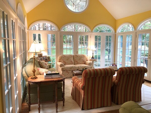 Relax in a spacious living room with a high ceiling, comfortable seating area, and large windows offering a view of the surrounding greenery.